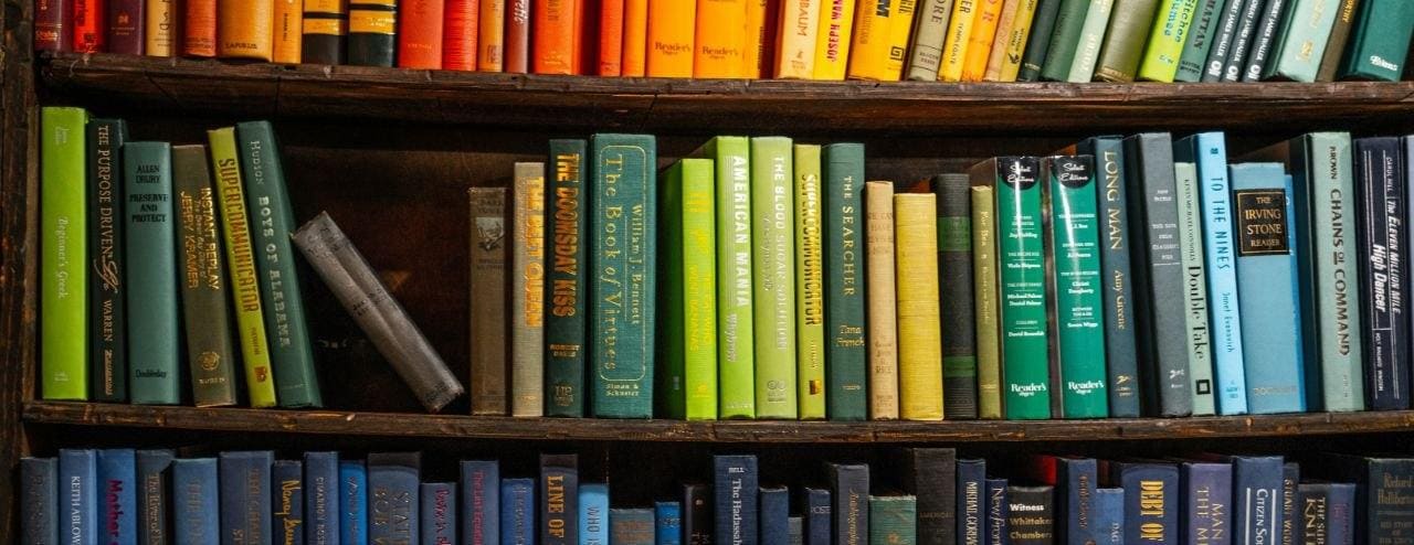 Dreireihiges Bücherregal mit bunten Buchrücken — oben Orange/Rot, mittig Grün/Gelb, unten Blau